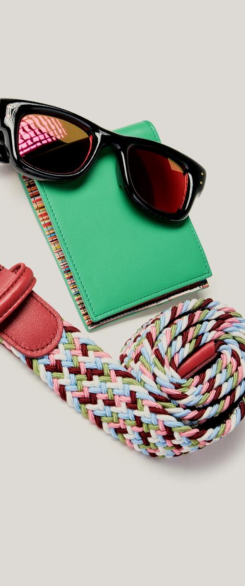 Lunettes de soleil à monture noire posées sur un portefeuille vert aux bords tissés multicolores, à côté d'une ceinture tressée multicolore enroulée avec une boucle en cuir rouge.