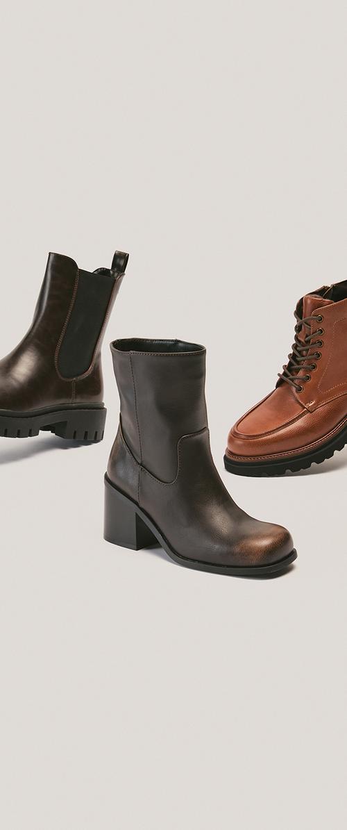 Tres botas de cuero: bota Chelsea marrón oscuro con suela gruesa, bota negra de tacón medio y bota corta con cordones color tostado sobre fondo liso.