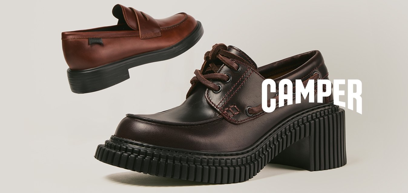 Dvoje hnědých kožených bot s černou robustní podrážkou, jedny mokasíny a jedny na šněrování, vystavené na čistém béžovém pozadí.