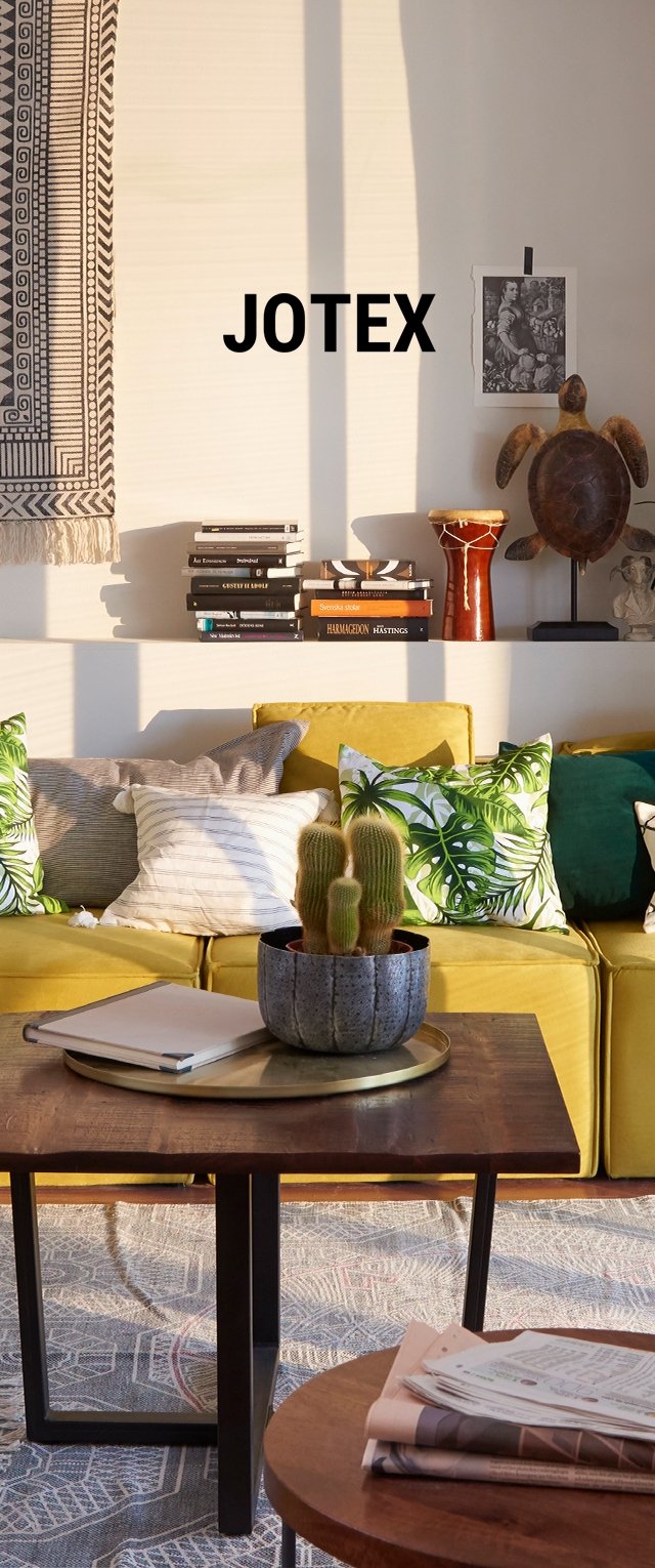 Sofá amarillo con almohadas decorativas, mesa de café de madera, planta decorativa y libros en una estantería, todo ello contra una pared clara y una alfombra estampada.