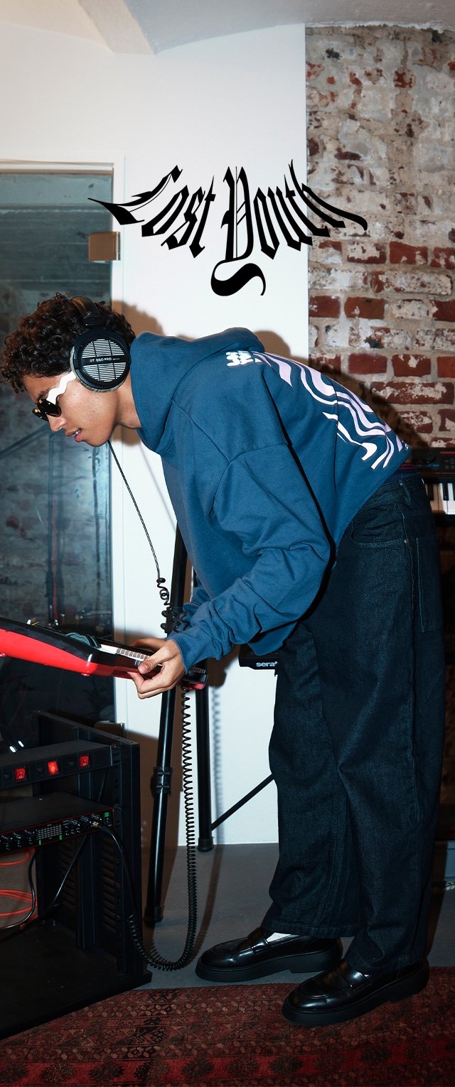 Persoon in een blauwe hoodie en koptelefoon die voorover buigt over elektronische muziekapparatuur in een kamer met een blootliggende bakstenen muur en vervormde tekst "Lost Youth" erboven.