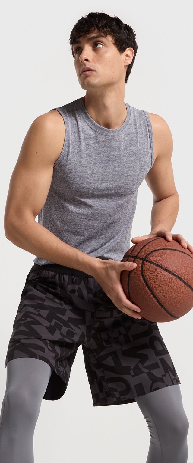 Camiseta atlética sin mangas de color gris, pantalones cortos negros con un patrón abstracto en gris oscuro y mallas compresivas grises claras. Sosteniendo un balón de baloncesto.