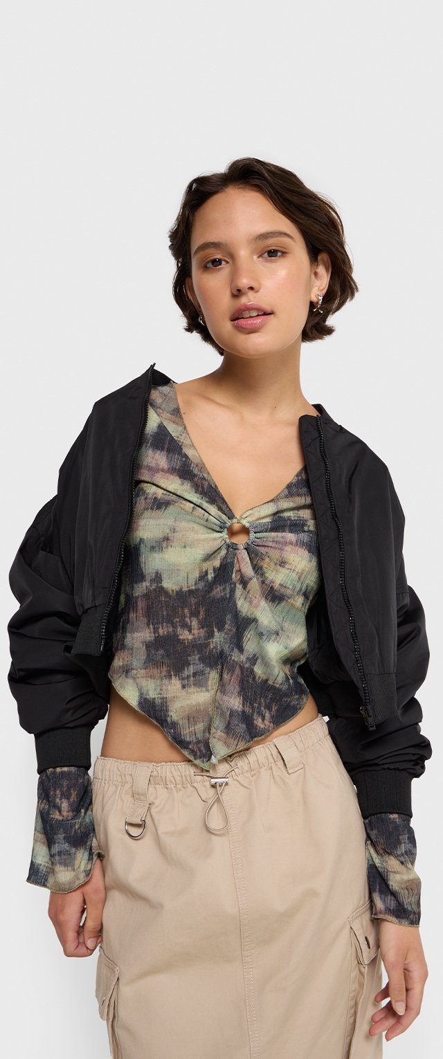 Czarna kurtka bomberka na wielokolorowej bluzce z wiązaniem na przodzie i okrągłym akcentem. Beżowa spódnica z troczkami. Skupienie na teksturach i wzorach.