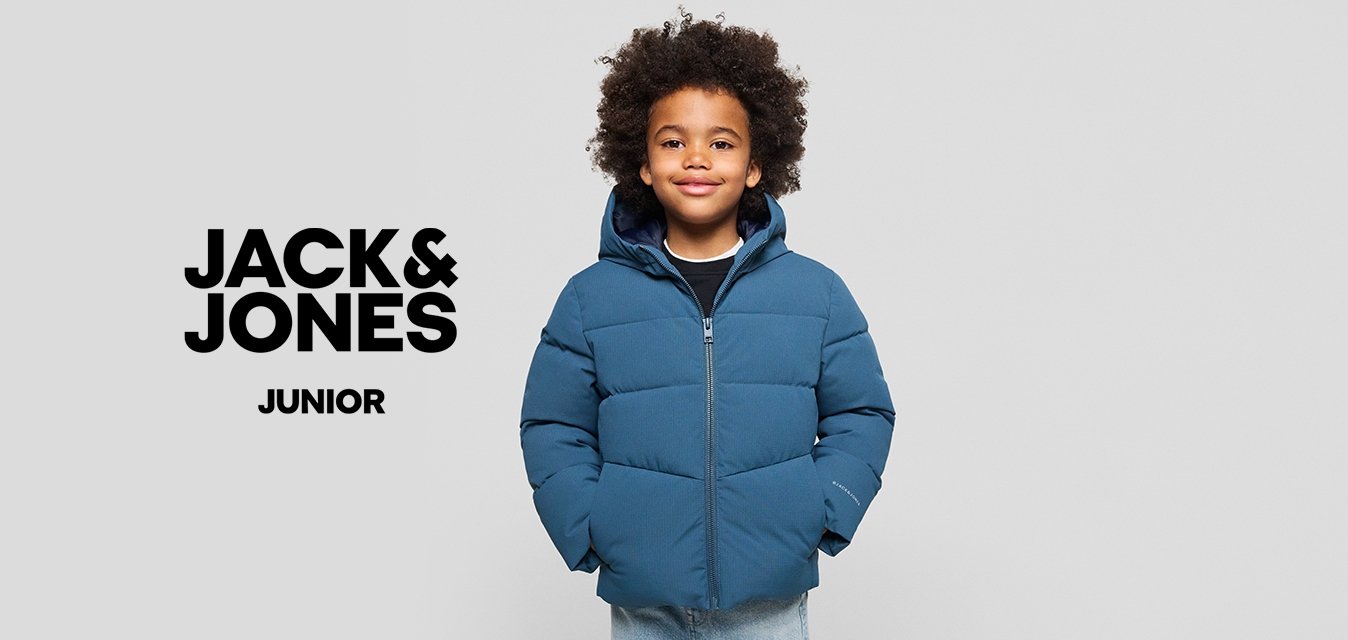 Mladé dítě s kudrnatými vlasy, oblečené v modré péřovce a světlých džínách, stojící s rukama v kapsách na jednobarevném pozadí.