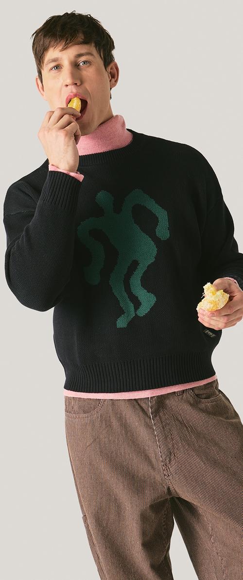 Jonge man in een zwarte trui met een groene abstracte figuur en een roze coltrui, die met beide handen een partje sinaasappel eet.