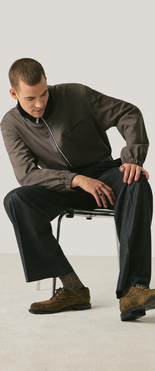 Chaqueta marrón con cremallera y cuello alto, pantalones oscuros y zapatos de ante marrones. Lleva calcetines estampados y está sentado en una silla contra un fondo claro.