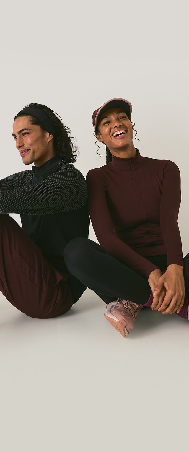 Deux individus sont assis côte à côte. L'un porte un haut noir zippé avec des épaules texturées et un pantalon sombre. L'autre porte un haut bordeaux et un pantalon assorti, avec une casquette. Les deux portent des chaussures de sport.