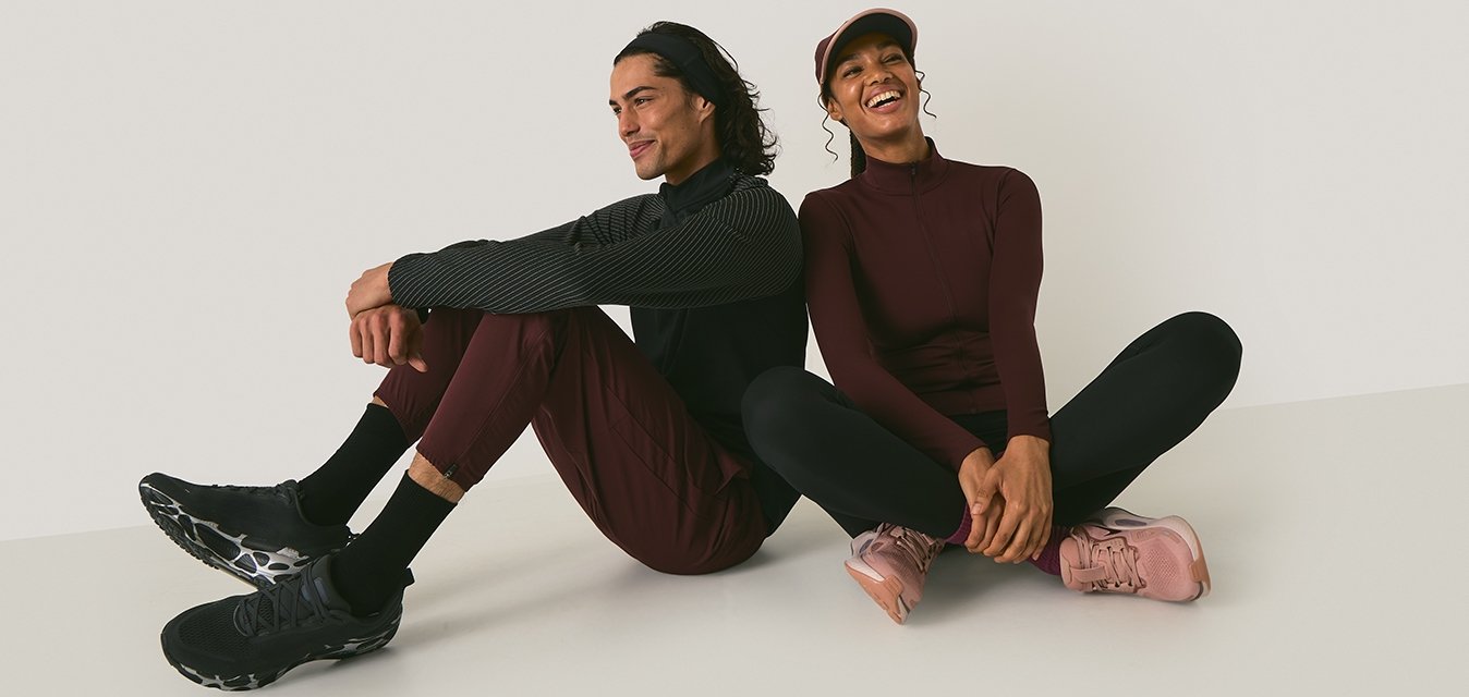 Dva jedinci sedí na světlém pozadí. Muž má na sobě černé strukturované tričko, vínové kalhoty a černé boty. Žena má na sobě vínovou mikinu na zip, černé legíny a růžové boty.
