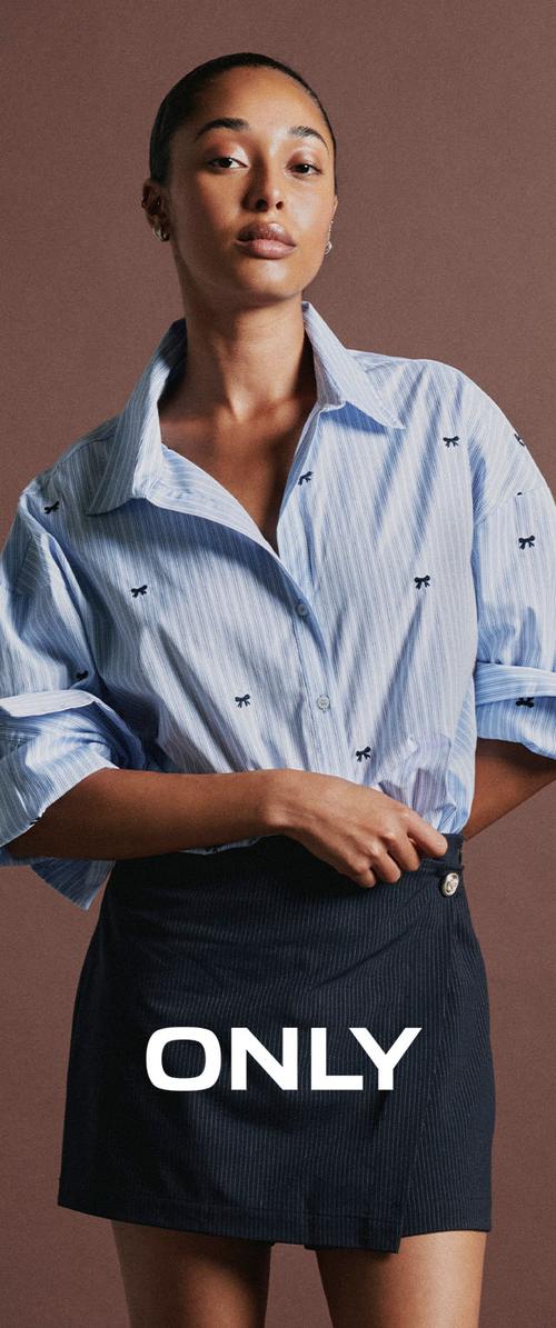 Une femme se tient devant un fond marron, portant une chemise à motifs bleu clair et une jupe foncée. Le mot ONLY apparaît en lettres blanches et grasses sur sa jupe.