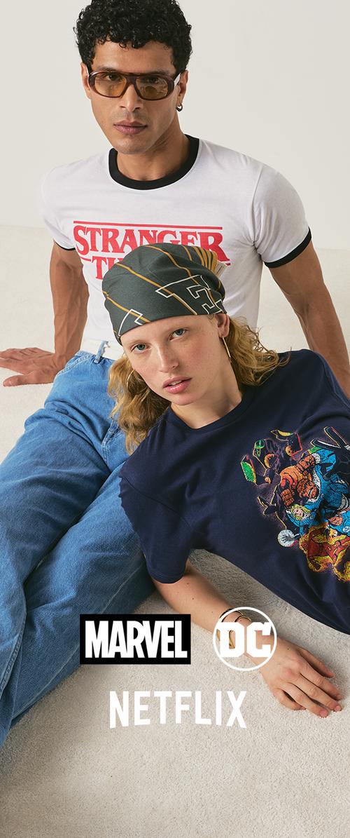 Two young adults, one wearing a Stranger Things t-shirt and sunglasses, the other in a Marvel comics t-shirt and headscarf, sitting on floor.
