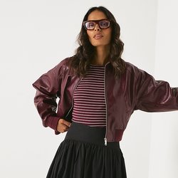 Burgundská kožená bomber bunda, pruhovaná halenka, černá plisovaná sukně, vysoké černé platformové boty po kolena. Model pózuje u bílé zdi.