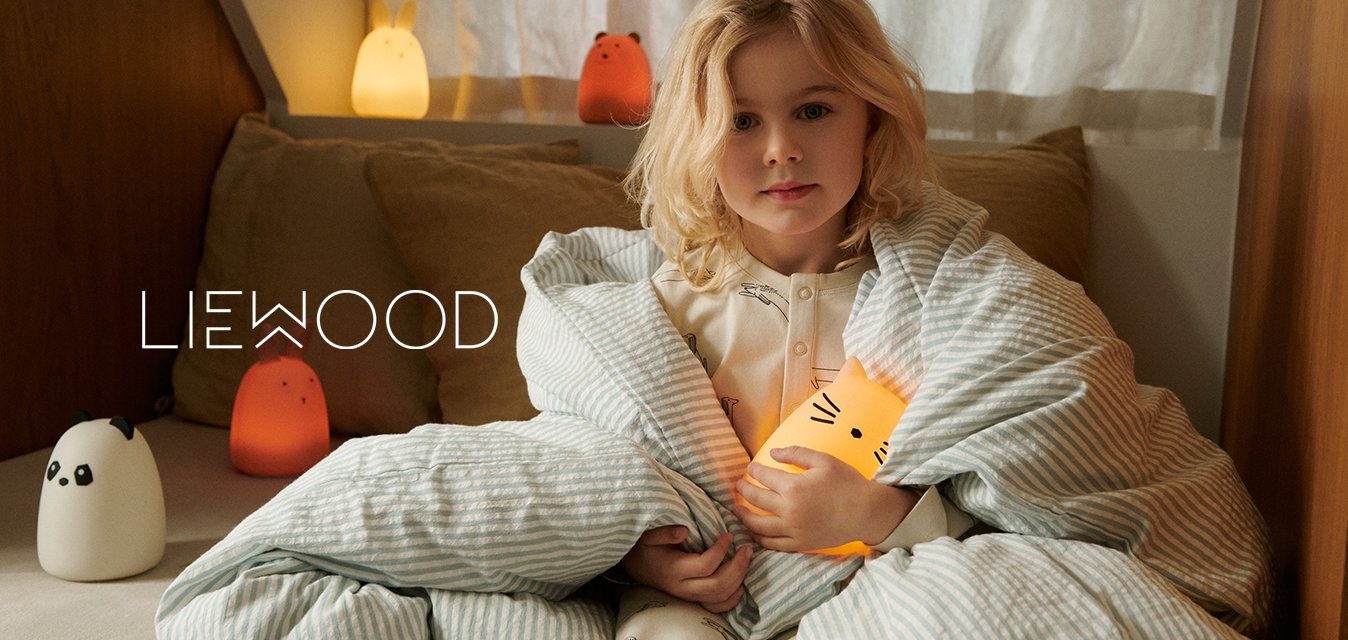 Dítě zabalené v světle modré pruhované dece drží měkkou, zářící lampu ve tvaru kočky. Viditelné jsou také měkké polštáře a lampy ve tvaru zvířat v teplých barvách.