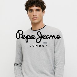 Mladý muž s kudrnatými vlasy, oblečený v světle šedém svetru a modrých džínách, stojící před jednobarevným bílým pozadím s jednou rukou v kapse.