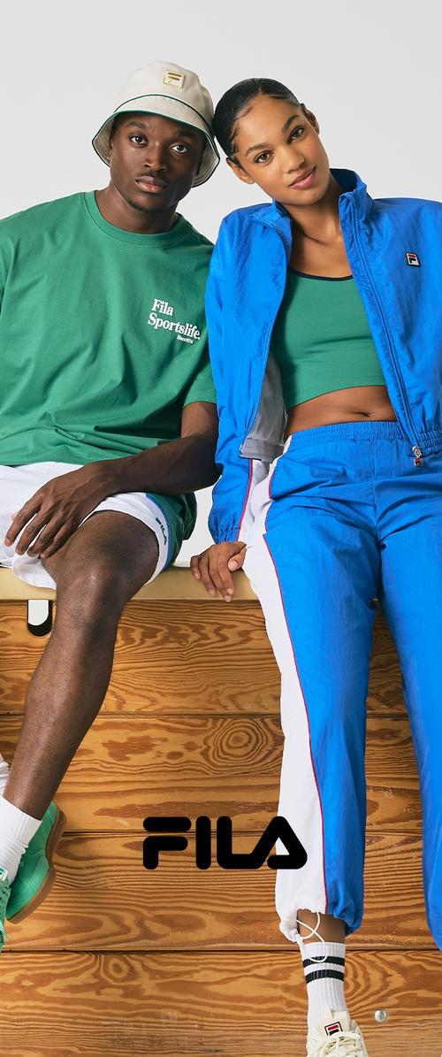 Joven con camiseta verde de FILA y pantalones cortos blancos, con gorra, y joven mujer con chándal azul de FILA y top verde, sentados en un banco de madera.