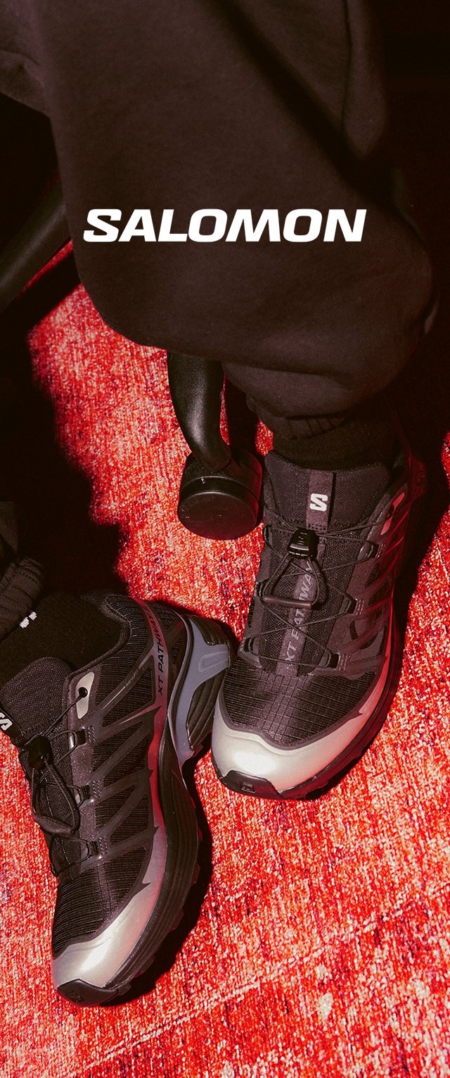 Las zapatillas de senderismo Salomon negras presentan un upper texturizado, acentos reflectantes y una suela gruesa y acolchada. Colocadas sobre un fondo de alfombra roja.