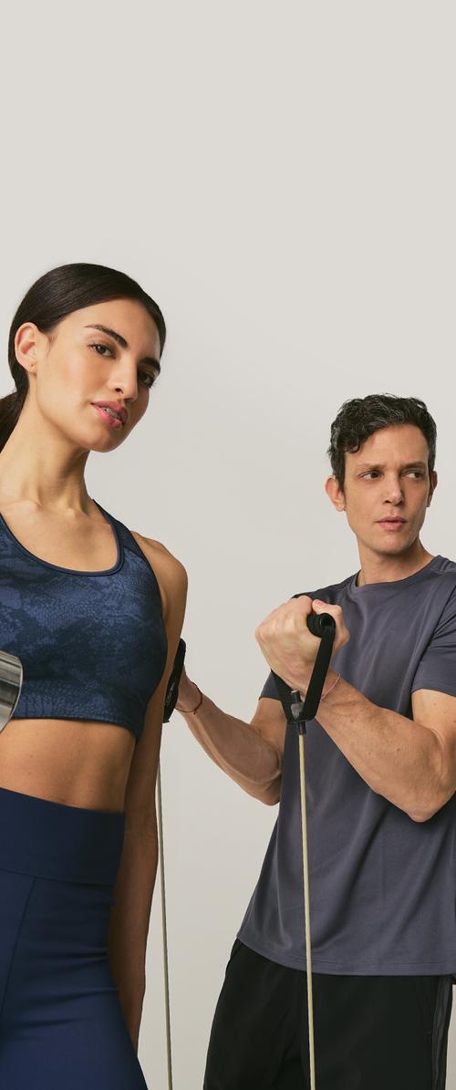 Dos personas con ropa deportiva: una con un sujetador deportivo azul oscuro y leggings de cintura alta, la otra con una camiseta gris. Ambas sostienen bandas de resistencia.
