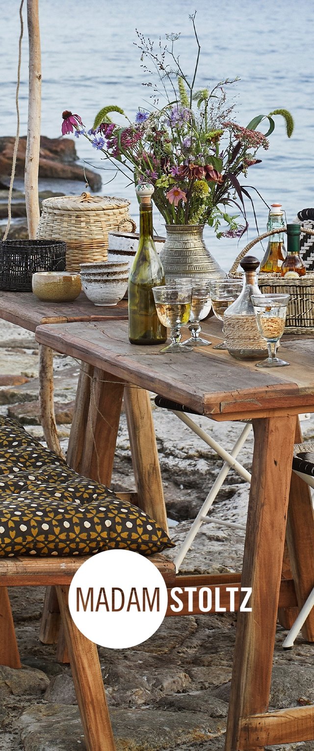 Wooden table with various glassware, a floral arrangement, patterned cushions, and woven baskets, set by a body of water.