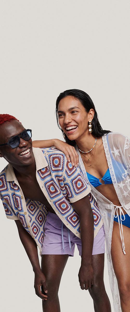 Deux personnes souriantes en vêtements d'été, l'une portant une chemise à motifs et un short violet, l'autre un bikini bleu avec un paréo transparent, posent proches l'une de l'autre.