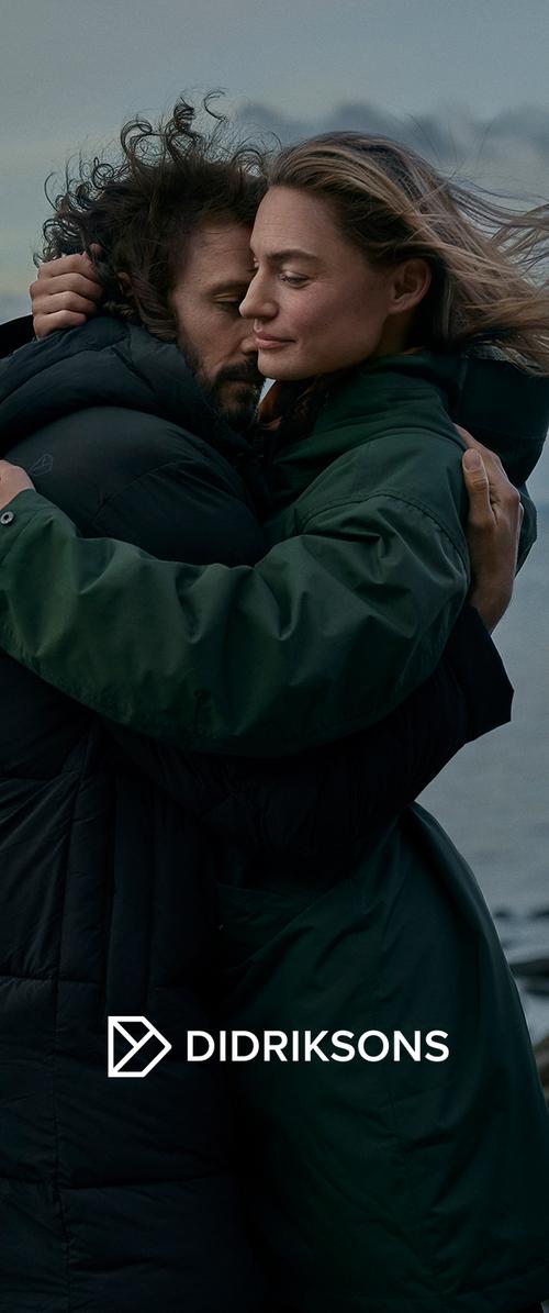 Dos personas se abrazan, vistiendo una chaqueta acolchada negra y un abrigo verde oscuro. La escena es al aire libre, con un cielo tenue y agua de fondo.