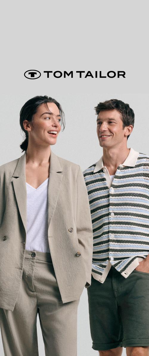 Frau im beigen Anzug und weißem Top und Mann im gestreiften Strickshirt und dunklen Shorts stehen lächelnd unter dem Tom Tailor-Logo vor grauem Hintergrund.