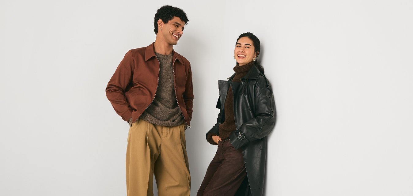 Dva lidé v módních podzimních oblečeních se usmívají a opírají se o jednoduchou bílou rohovou zeď v minimalistické místnosti.