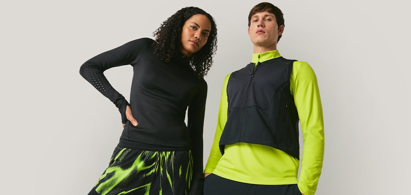 Dva modelové stojí na jednobarevném pozadí, jeden má na sobě černé a neonově zelené sportovní oblečení, druhý černý top a neonově zelenou bundu s vestou.