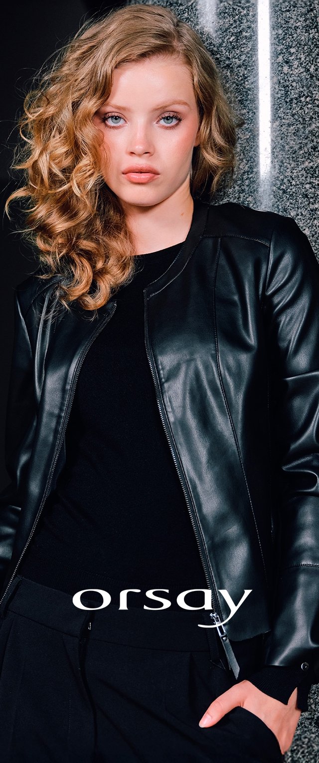 Mujer con cabello rizado rubio que lleva una chaqueta de cuero negra y un conjunto negro, de pie frente a una pared gris texturizada con una tira vertical de luz.