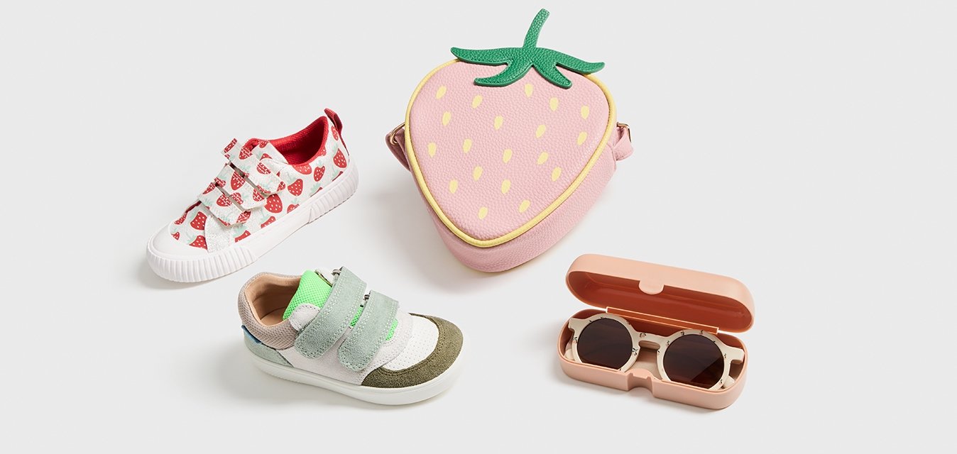 Bílé boty s červenými vzory jahod, zelené a šedé boty na suchý zip, růžová taška ve tvaru jahody a růžový pouzdro na sluneční brýle.