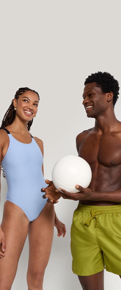 Zwei Personen stehen zusammen: eine in einem hellblauen Badeanzug, die andere in grünen Badeshorts, beide halten einen weißen Volleyball.