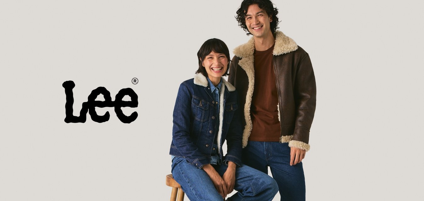 Dva lidé ve džínových a ovčích bundách; jeden sedí na stoličce, zatímco druhý stojí, oba se usmívají na jednobarevném pozadí.