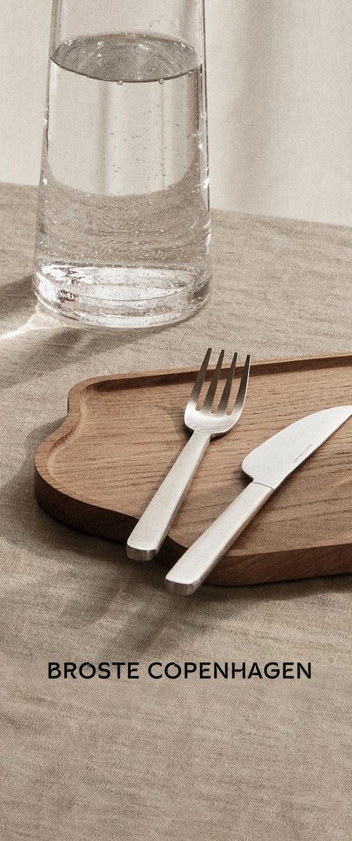 Plateau en bois aux bords incurvés contenant une fourchette et un couteau en acier inoxydable. En arrière-plan, un verre d'eau et une nappe beige texturée.