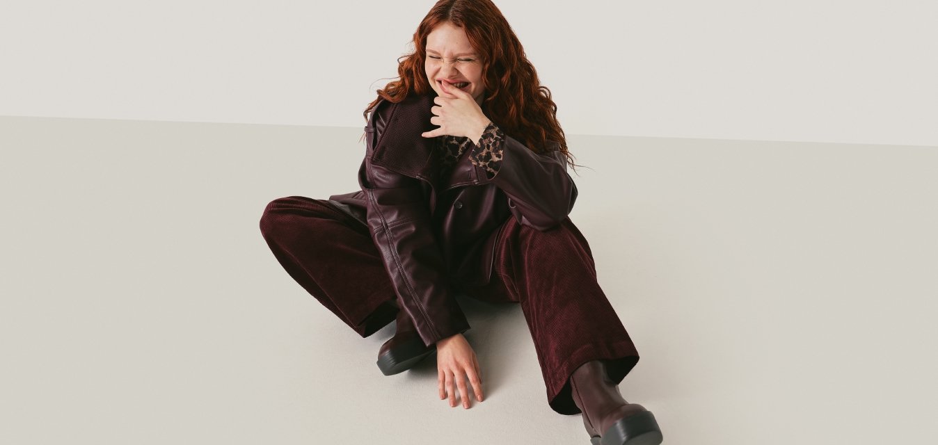 Mladá žena se zvlněnými rudými vlasy sedí na podlaze v tmavě fialových kalhotách a bundě, usmívá se a dotýká se tváře na neutrálním pozadí.