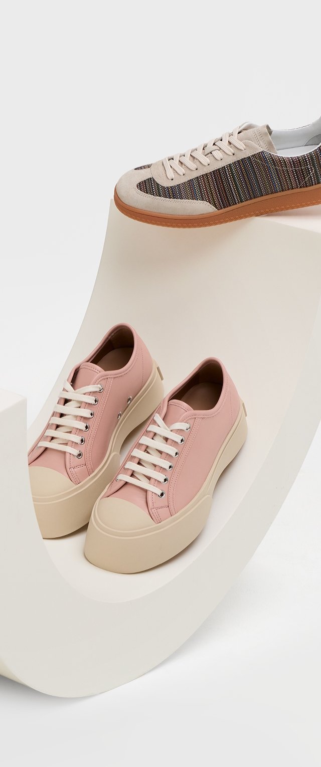 Ljusrosa sneakers med en slät yta, vita snören och beige gummisulor. Det andra paret har en strukturerad, flerfärgad randdesign.