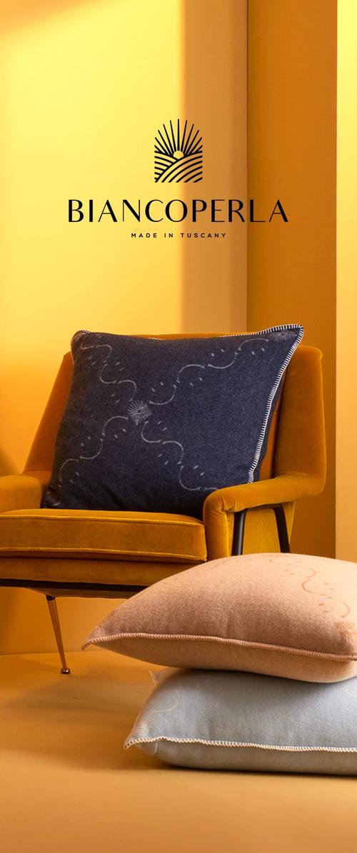 Cojín azul oscuro con bordado claro, apoyado sobre un cojín azul claro y uno color beige, frente a un fondo amarillo y una silla.
