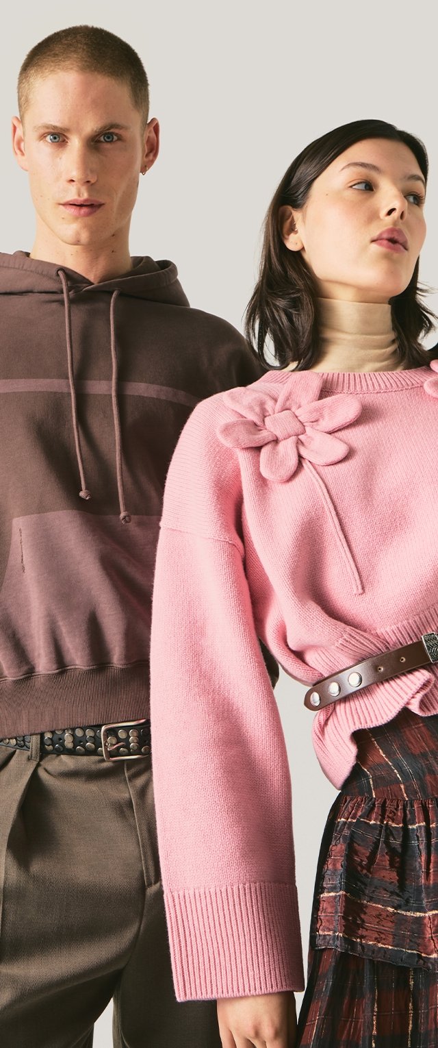 Joven con sudadera marrón con capucha y pantalones con cinturón tachonado, de pie junto a una mujer con suéter rosa con detalle floral, cuello alto, cinturón y falda estampada.