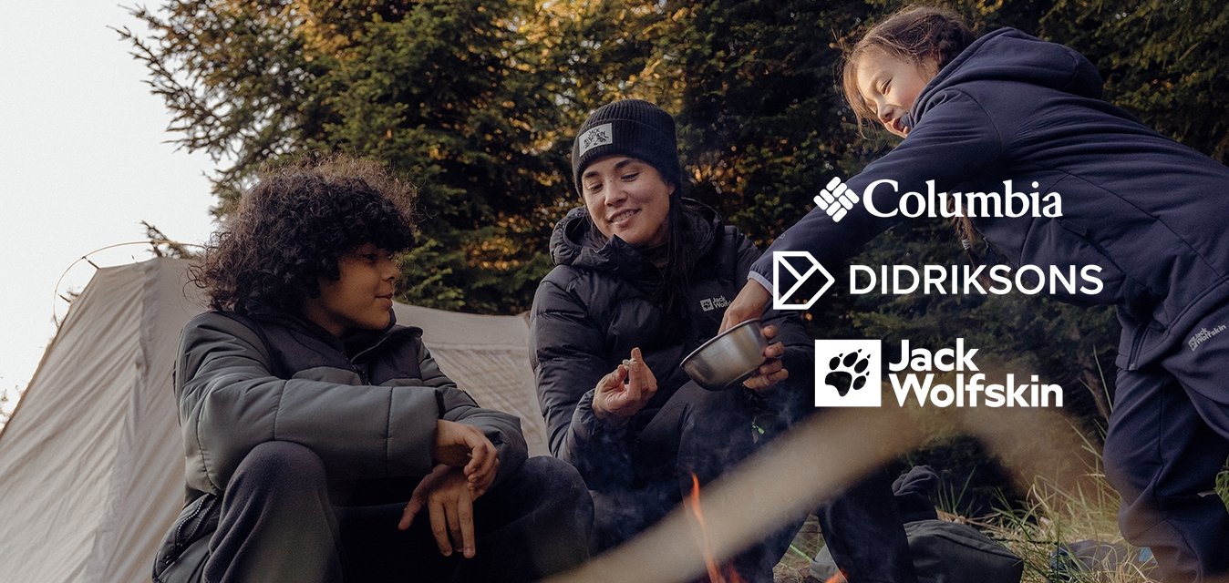 Tři lidé u ohniště v lese, oblečeni v tmavých bundách a botách. V pozadí je vidět stan, stromy a tráva.