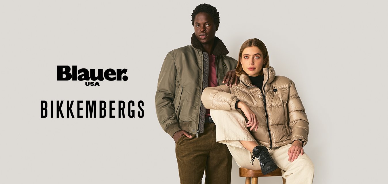 Dva dospělí mají na sobě teplé bundy: jeden stojí v olivovém bomberu a hnědých kalhotách, druhý sedí na stoličce v béžovém pufru a krémových kalhotách.