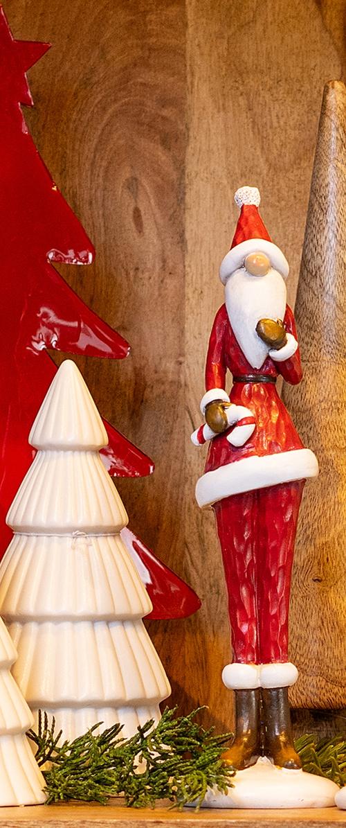 Keramische kerstmanfiguur in rood met witte accenten, die een geschenk vasthoudt; staand naast witte en rode boomdecoraties op een houten achtergrond.