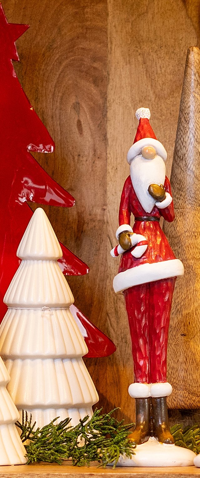 Figura de cerámica de Santa en rojo con detalles en blanco, sosteniendo un regalo; de pie junto a decoraciones de árbol en blanco y rojo sobre un fondo de madera.