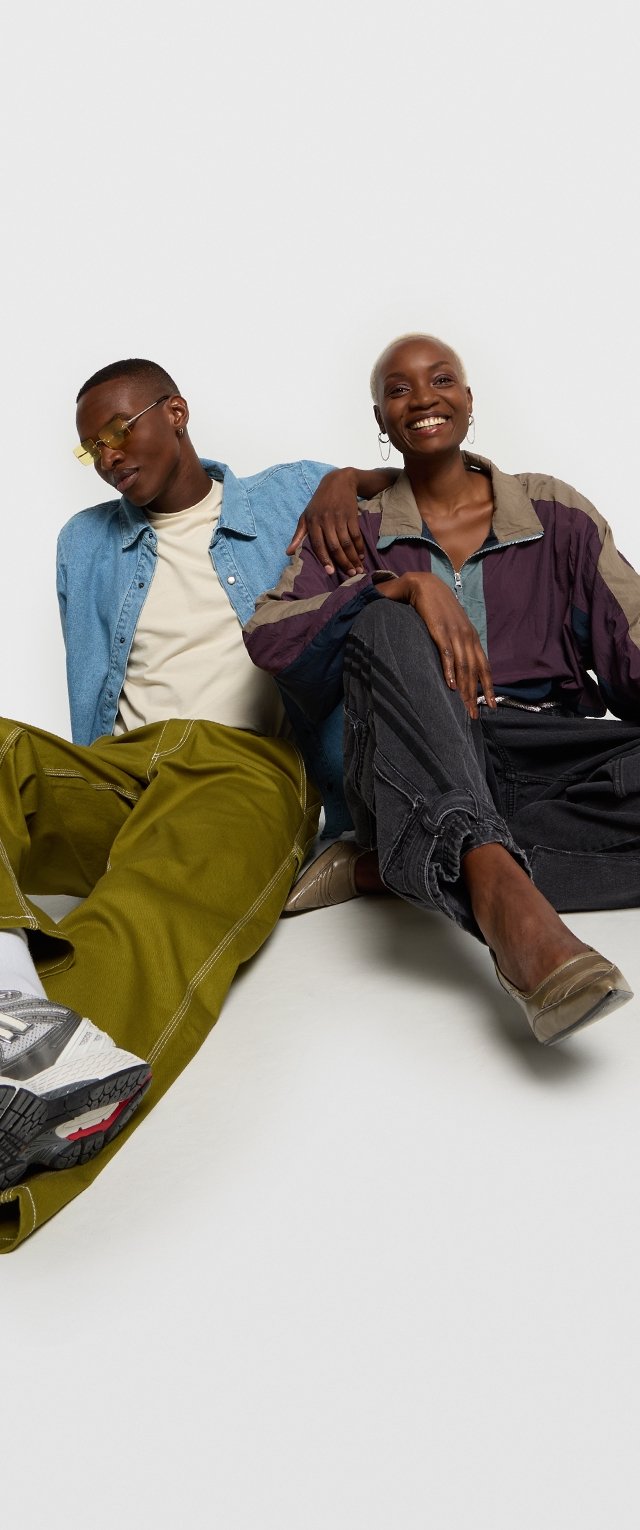 Dos personas están sentadas de manera informal, vistiendo una camisa de mezclilla, una chaqueta multicolor, pantalones verdes y zapatillas modernas, frente a un fondo liso.