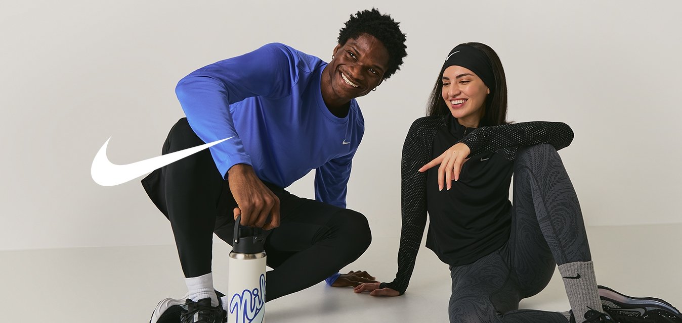 Dva lidé v atletickém oblečení sedí na podlaze a usmívají se; jeden drží láhev s vodou, oba vypadají uvolněně a přátelsky.