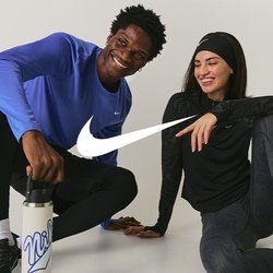 Dva lidé v atletickém oblečení sedí na podlaze a usmívají se; jeden drží láhev s vodou, oba vypadají uvolněně a přátelsky.