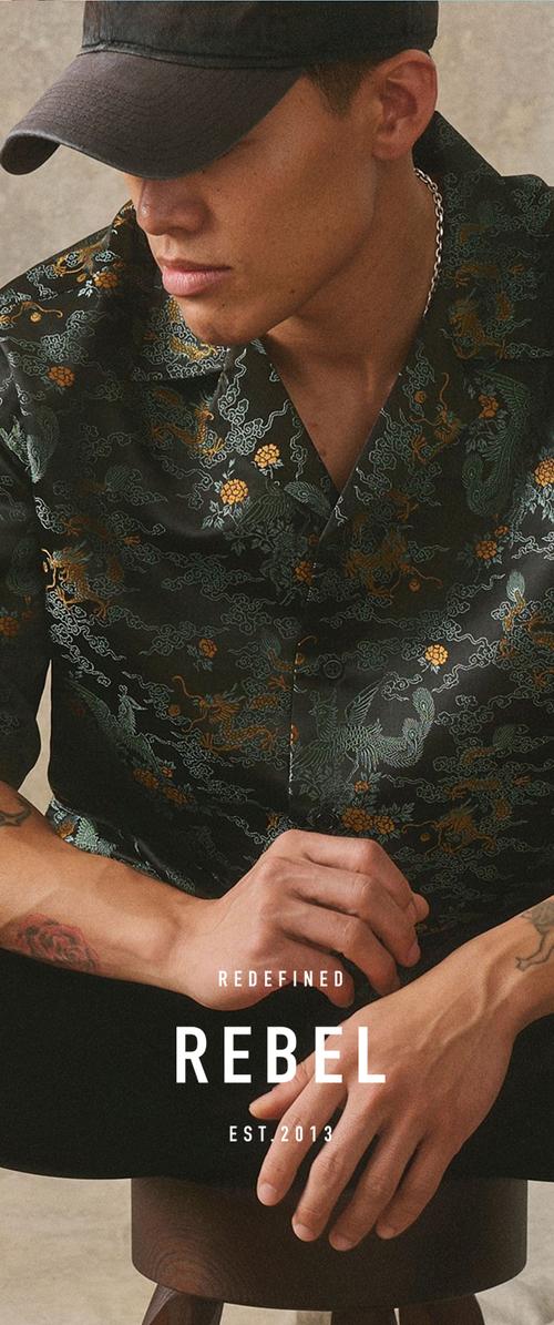 Junger Mann mit schwarzer Kappe und dunklem, gemustertem Hemd mit Drachen und Blumen, sitzt und legt die Hände auf die Knie.