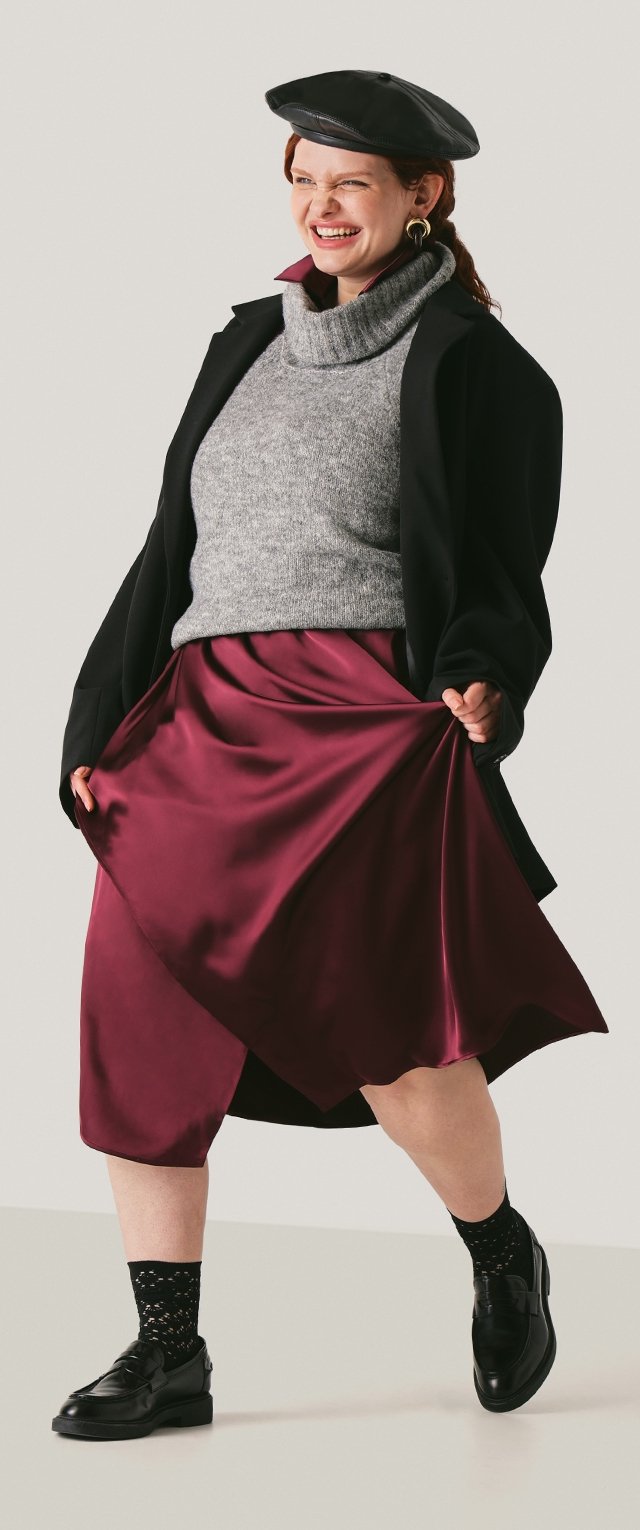 Modell trägt einen schwarzen Mantel über einem grauen Strickpullover und einem bordeauxroten Satinenen Rock, kombiniert mit gemusterten schwarzen Socken und schwarzen Schuhen.