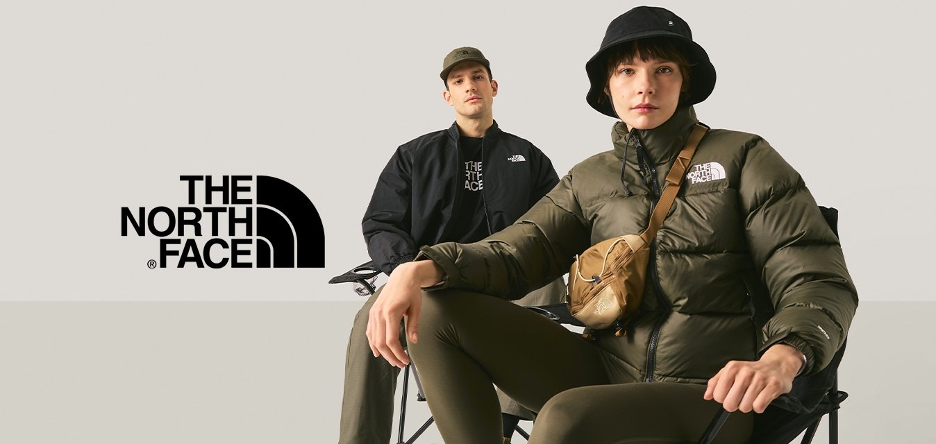 Dva mladí dospělí sedící na skládacích židlích na jednobarevném světle pozadí, jeden má na sobě černý klobouk typu bucket a prošívanou bundu.