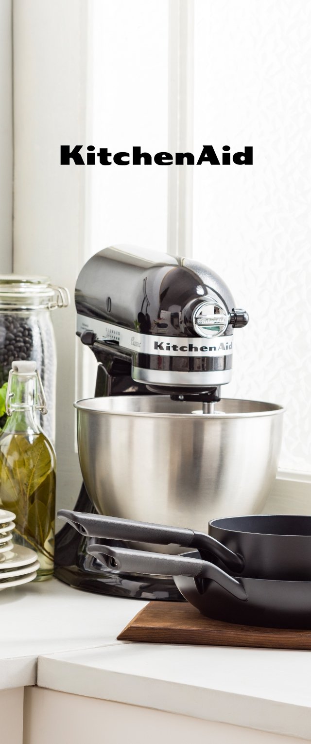 Schwarzer KitchenAid-Küchenmixer mit Edelstahlschüssel auf der Küchenarbeitsplatte, neben Glasflaschen, Tellern und gestapelten schwarzen Pfannen auf einem Holzbrett.