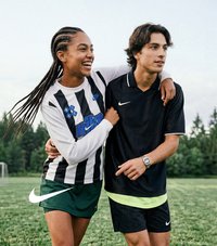 Zwei junge Menschen in Sportbekleidung lächeln und gehen über ein grasbedecktes Feld mit Bäumen im Hintergrund.