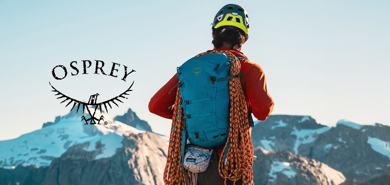 Modrý batoh s texturovaným povrchem, na kterém jsou připevněny oranžové horolezecké laná, nošený přes oranžovou košili s dlouhým rukávem, na pozadí hor.