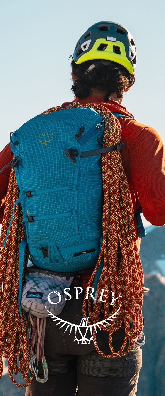 Niebieski plecak wspinaczkowy z siatkowymi wzorami, liczne paski i przymocowane czerwono-żółte liny wspinaczkowe. Osoba nosząca kask i sprzęt wspinaczkowy.