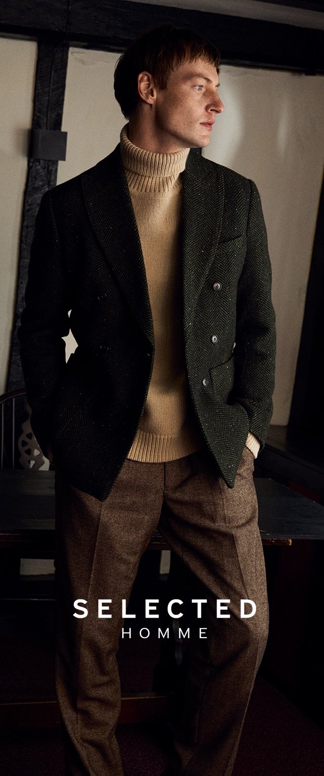 Veste double boutonnage en tweed à chevrons verts sur un col roulé beige, associée à un pantalon texturé marron. Le modèle pose devant un fond neutre.